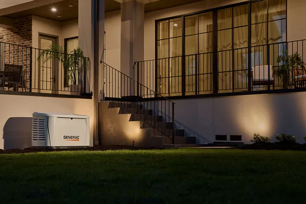 Modern home illuminated at night, featuring an outdoor balcony and staircase with black railings, large glass windows with curtains, and a Generac generator installed on the lawn beside the house.